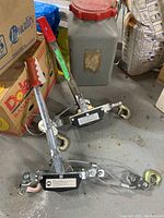 Photo of two cable winches with red-handled grips, steel cables, and hooks lying on a concrete floor near various boxes and a metal container.