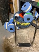Photo showing two blue and white styrofoam hand weights next to a blue bag labeled Rocker Stopper and the stainless steel two-step ladder leaning nearby.