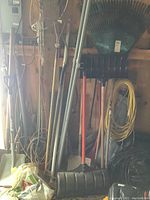 Overall view of rakes, shovels, and snow shovels leaning against wall including metal green leaf rake and black plastic snow shovels.