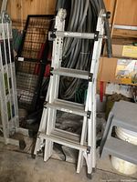 Aluminum scaffolding ladder folded and standing upright in a garage area with nearby tools and materials.