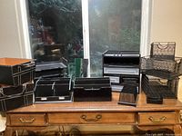 Photo showing full collection of black organizational office totes, racks, bins, and drawer organizers arranged on wooden surface in front of window.