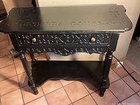 Front view of carved black console table showing detailed top edge carvings, pull-out drawer, and lower shelf with curved edges.