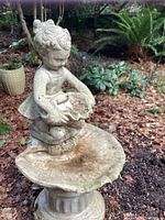 Close-up photo of the entire fountain showing the girl holding a shell and bath basin below on pedestal with garden background.