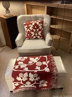 Cream wingback chair with red patterned pillow, cream rectangular storage bench with red and white throw blanket draped on top, carpeted room background.