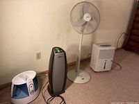 Wide shot showing all four appliances lined up against a wall: blue and white Vicks humidifier, black and silver Hunter tower air purifier, white Windmere stand fan, and white Energy Star dehumidifier.