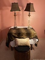 Front view of the two table lamps with leopard print shades, small leopard print vanity chair with black seat and storage, beige throw pillow, and leopard print blanket draped on a table.
