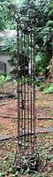 Full view of the tall metal yard art tower standing outdoors on grass, showing cylindrical wireframe design with decorative top and solar string lights wrapped around.