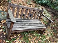 Front view of the weathered wooden outdoor bench showing slatted seat and backrest with armrests, surrounded by fallen leaves outdoors.