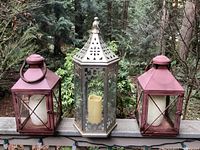 Three patio lanterns with candles on a railing: two red metal with large candles and one silver metal with decorative cut-outs and LED candles.