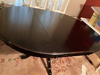 Side angle showing round black wood dining table with split top expanded by leaf, pedestal base with four legs visible