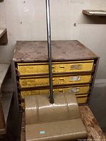 Photo of the top and front of the vintage metal tool box with four yellow drawers marked Mechanics Choice; metal dustpan with handle resting in front.
