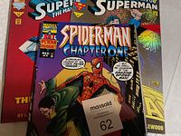 Three comic books stacked with the top one showing Spider-Man Chapter One, issue 1, Marvel Comics. Below it are two Superman comic books.