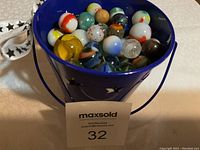 Blue metal bucket filled with assorted glass marbles including various colors and patterns with handle visible.