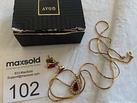 View of Avon box, necklace chain, pendant with red and clear stones, and matching earrings arranged on a white surface.