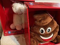 Photo showing the brown smiling poop emoji plush Bluetooth speaker and part of the white unicorn plush Bluetooth speaker, both in branded red boxes.