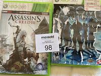 Front view of Assassin's Creed III and NBA 2K13 Xbox 360 game cases with auction card in center.