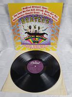 The Beatles Magical Mystery Tour vinyl LP record with purple Capitol label on cream sleeve, front cover leaning behind with colorful psychedelic artwork.