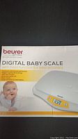 Box of Beurer BY80 digital baby scale showing product image, baby photo, and features like hold function and automatic switch off.