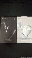 Two Cambridge Audio Melomania 1+ earbud boxes shown side by side, one black box, one white box, showcasing the product names and images.