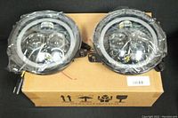 Front view of two round Jeep Wrangler JL headlights in plastic wrap, showing clear lenses and LED clusters inside, resting on cardboard box.