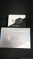 Two LED projector boxes stacked, one with image of compact white and black LED projector, the other an illustrated video projector with LED source.