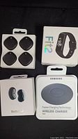 Photo showing all items together including Samsung Galaxy Fit2, wireless charger pad, Galaxy Buds Live box, and four Samsung AirTags in packaging.