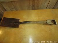 Full length image of the antique shovel laid flat on a wooden table, showing the metal blade and wooden handle with D-grip.