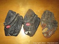 Three worn baseball gloves side by side on a flat surface; two black Rawlings gloves with red logos and one brown glove with adjustable strap.