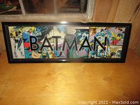 Framed Batman comic strip wall art showing word BATMAN and vintage comic panels, on wooden surface with window background