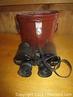 Black Celestron binoculars with focus wheel and zoom markings, placed in front of brown leather carrying case on wooden surface.