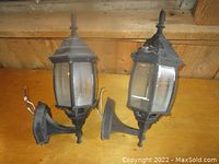 Two black metal exterior coach lights with clear glass panels shown side by side on a wooden surface.