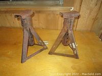 Pair of rusted metal jack stands viewed from front on wooden surface, showing triangular bases and adjustable arms.