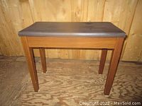 Front view of wooden piano bench with rectangular padded seat on wooden legs against wooden wall and floor.
