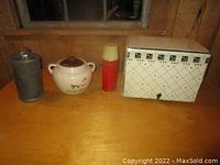 Four vintage kitchen items on wooden table under window: metal mixer/churn, cow cookie jar, red thermos, white metal bread box with cow pattern