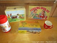 Photo showing a vintage Lassie metal lunch box, Korking Spool Kit box, Howdy Doody set box, Disney thermos, and a red Tippy child mug on a wooden surface.