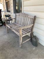 Weathered teak bench placed on a concrete porch, showing full front and side view including carved details on the upper backrest and bottom front panel.
