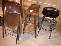 Full view of all three seating items against an unfinished basement wall: metal folding chair, wooden stool, and Snap-On racing stool.