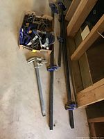 Photo showing multiple pipe clamps of varying length lying on the floor next to wall studs, along with a box containing many smaller clamps.