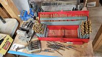 View of multiple red drill bit cases with assorted drill bits arranged by size, two metal vises behind, loose bits and tips on the surface.