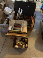Overview photo showing boxes containing easel, paints, foam boards, blank CDs, rolls of tape, building blocks, and wire components in basement setting