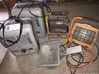 Two Patton heaters in gray color with power cords, and three different work lights, including a large floodlight and two caged cage lights on stands, all placed on a basement floor.