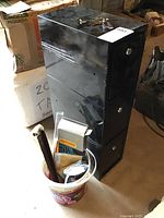Black metal locking cabinet with three lockable compartments, placed on a concrete floor with a bucket of cleaning supplies beside it.