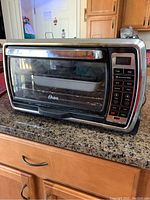 Front view of Oster toaster and convection oven showing glass door, handle, and digital control panel with multiple functional buttons.