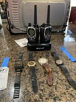 Front view of four watches laid in a row with Midland walkie talkie set charging base behind them, placed on a speckled countertop.