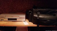 Silver Sylvania VCR alongside black camcorder carrying bag on wooden shelf