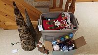 Photo showing two light-up cone Christmas trees next to a box with assorted ornaments and a plastic bin full of stockings and holiday items.