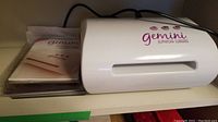 Photo showing the white Gemini Junior die cutting and embossing machine with the power cord bundled on top. Machine body is plain with pink Gemini Junior branding on front.