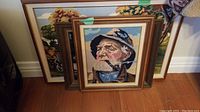Three framed needlepoint pieces stacked against a wall including a portrait of an elderly man and landscape scenes.