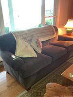 Front view of dark gray corduroy three-seater sofa with seat cushions, two decorative pillows and fleece blanket draped over back near a window and lamp