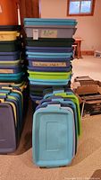 Stack of 13 plastic storage bins with various colored lids including blue, green, gray, and purple, some labeled.